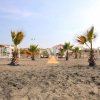 Spiaggia sabbiosa con palme, lettini e un sentiero in legno che conduce a costruzioni sullo sfondo.
