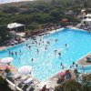 Piscina all'aperto con molte persone. Ombrelloni e lettini attorno. Vegetazione circostante.