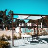 Area piscina con tavolo e panchina sotto un pergolato. Persone nuotano e si rilassano.
