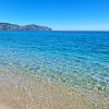 Mare cristallino e spiaggia sabbiosa con colline in lontananza sotto un cielo azzurro.