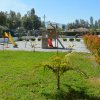 Un parco giochi con altalene e scivolo, circondato da un prato verde e arbusti rossi.