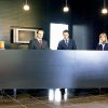 Tre persone in abiti formali dietro un bancone scuro, con luci pendenti e una pianta a sinistra.