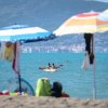 Persone in kayak sul mare, viste da una spiaggia con ombrelloni colorati.