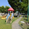 Parco giochi con scivolo colorato e altalene su prato verde, circondato da alberi.