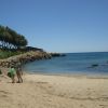 Tre persone camminano su una spiaggia sabbiosa verso il mare, con rocce e alberi sullo sfondo.