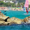 Spiaggia affollata con persone che fanno windsurf e nuotano vicino a rocce.