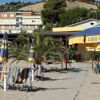 Spiaggia con sedie a sdraio e ombrelloni gialli e blu, palme e un edificio sullo sfondo.
