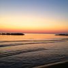Un tramonto sul mare con cielo arancione e blu, onde leggere e scogliere distanti.