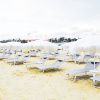 Spiaggia sabbiosa con ombrelloni bianchi e lettini grigi disposti in file ordinate.