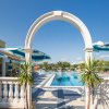 Ingresso ad arco con vista su piscina e ombrelloni, circondato da palme sotto un cielo azzurro.