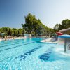 Piscina all'aperto con giochi d'acqua, ombrelloni blu e vegetazione attorno.