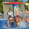 Bambini giocano sotto una cascata d'acqua in piscina, circondati da sdraio e ombrelloni.