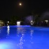 Una piscina illuminata di blu di notte con una fontana e la luna visibile nel cielo.