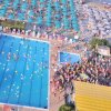 Piscina affollata in un complesso balneare con ombrelloni e lettini verdi e blu.