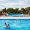 Una piscina con persone che nuotano, circondata da alberi e ombrelloni blu.