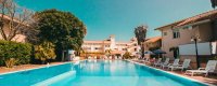 Piscina lunga con lettini bianchi ai lati e palme, circondata da edifici.