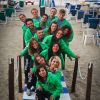 Gruppo di persone in felpe verdi, posano sorridenti su passerella blu e bianca sulla spiaggia.