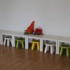 Tavoli bianchi e sgabelli colorati con due pupazzi soffici su una panca contro un muro bianco.