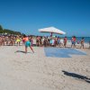 Gruppo di persone sulla spiaggia, alcune partecipano a un'attività sportiva sotto il sole.