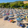 Spiaggia con ombrelloni gialli e blu, sedie a sdraio e vegetazione collinare sullo sfondo.