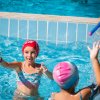 Bambini giocano in piscina con cappellini e tubi colorati, uno spruzza acqua.