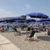 Spiaggia con ombrelloni blu e sedie a sdraio su ghiaia, mare in lontananza.