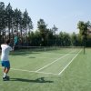 Un ragazzo gioca a tennis su un campo verde all'aperto, circondato da alberi.