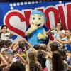Un grande pupazzo a forma di tartaruga blu intrattiene un gruppo di bambini sorridenti.