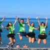 Gruppo di persone in magliette verdi che saltano in spiaggia davanti al mare.