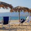 Spiaggia con lettini blu e ombrelloni di paglia, vista sul mare e montagne.