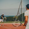 Due persone che giocano a tennis su un campo all'aperto vicino al mare, con una rete di recinzione.