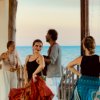 Donne danzano con gonne colorate vicino al mare mentre un uomo suona la chitarra al tramonto.