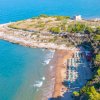 Veduta aerea di una spiaggia con ombrelloni, promontorio roccioso e mare blu.